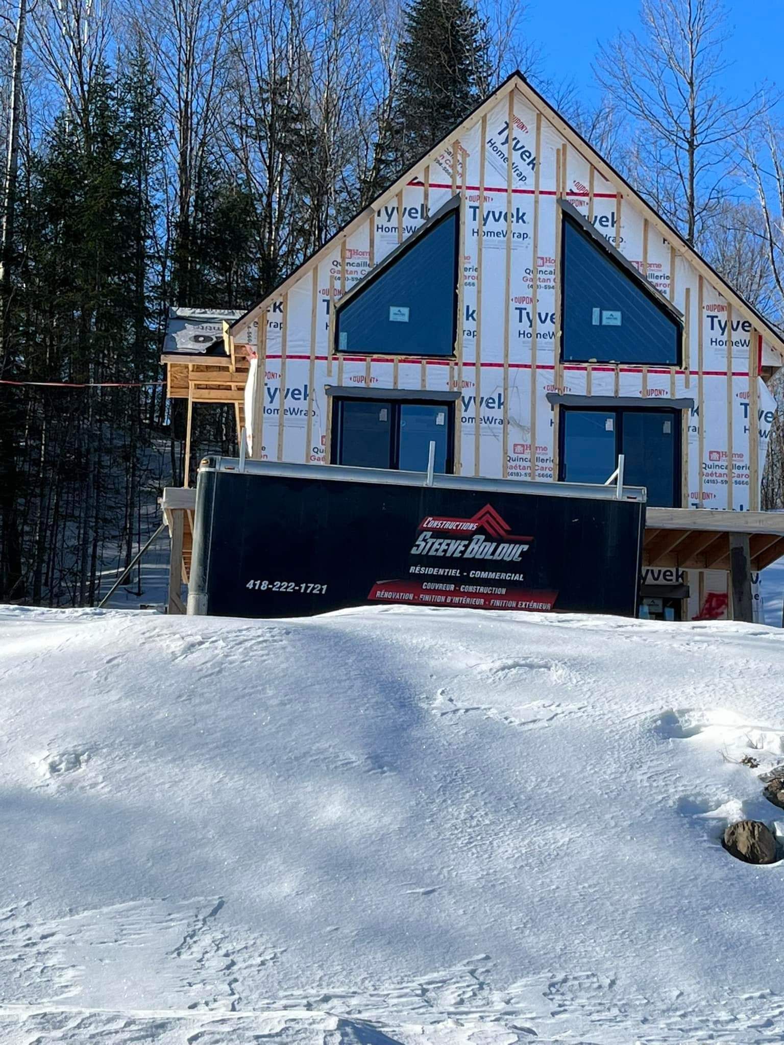 Maison en construction en hiver, sous la neige. Bâche noire devant. Grandes fenêtres.