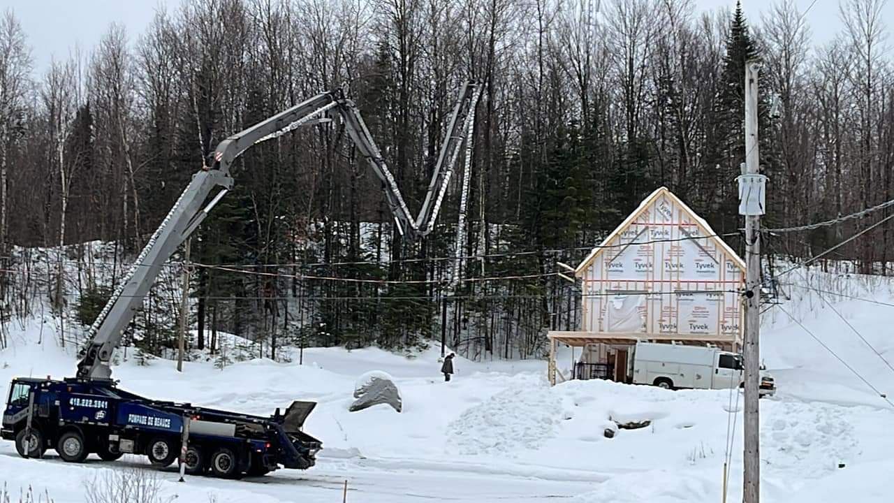 Un camion-pompe à béton déverse du béton dans la structure d'une nouvelle maison, dans un décor boisé et enneigé.