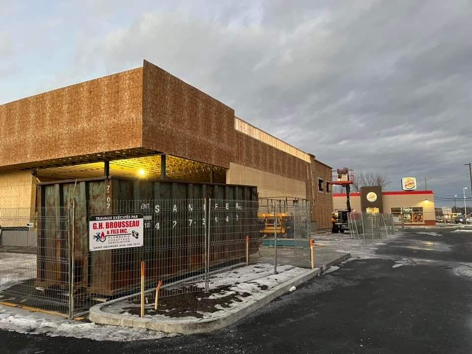 Chantier de construction avec un bâtiment partiellement recouvert de panneaux de bois, à côté d'un restaurant de restauration rapide en activité.