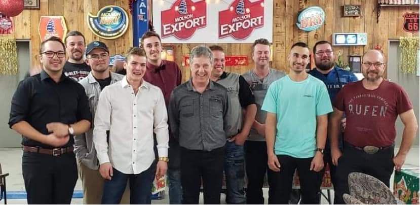 Groupe d'hommes posant ensemble pour une photo, en intérieur. Fond en panneaux de bois, enseignes de bar.