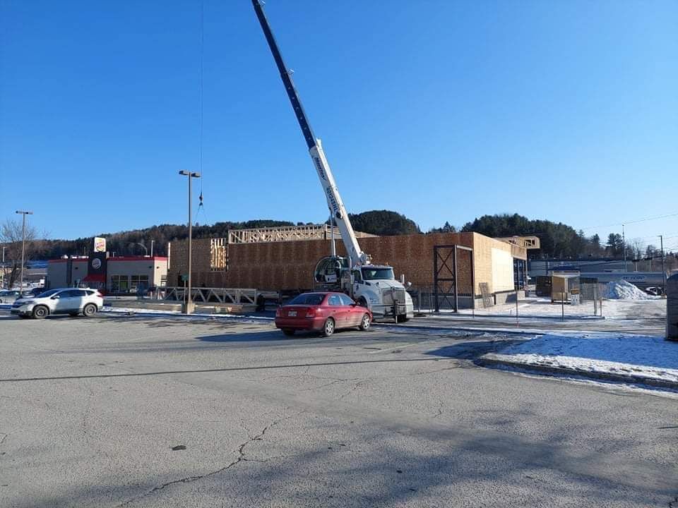Une grue soulève un panneau mural en bois sur un chantier de construction, sur un parking, par une journée ensoleillée.
