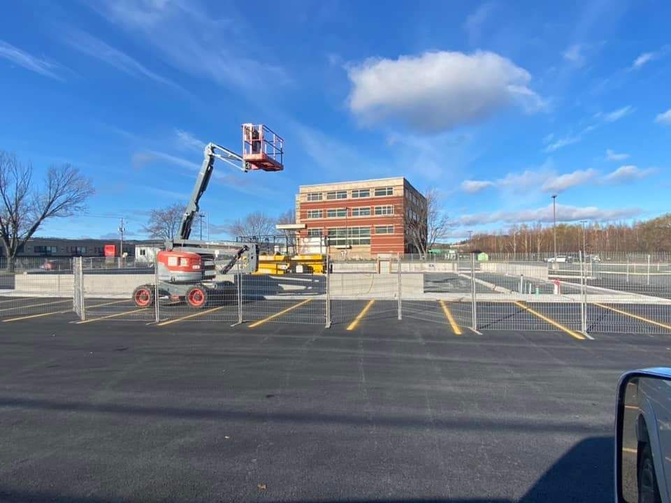 Chantier de construction avec une nacelle élévatrice orange, un bâtiment et un parking nouvellement goudronné avec des lignes jaunes sous un ciel bleu.