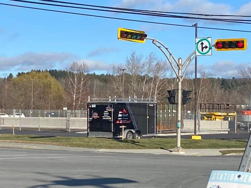Feux de circulation à une intersection, avec une remorque noire devant. Ciel bleu vif avec des arbres en arrière-plan.