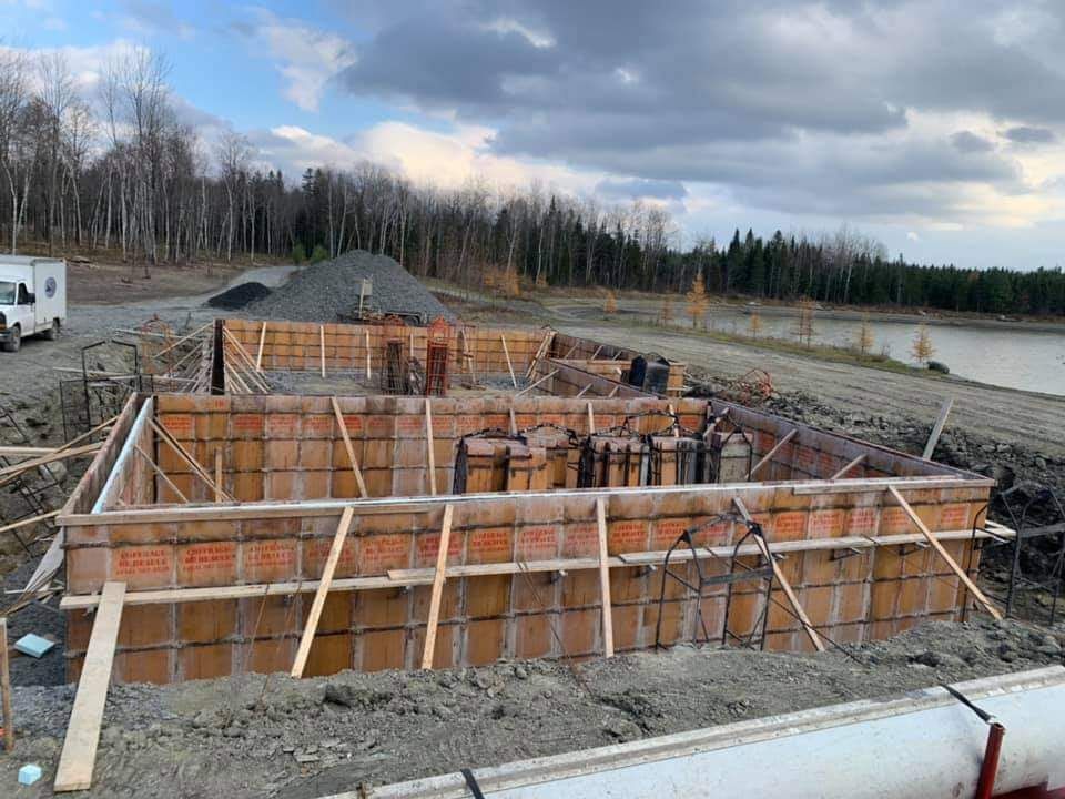 Chantier de construction, coffrages en bois pour les fondations d'un bâtiment ; terre, eau et arbres en arrière-plan.