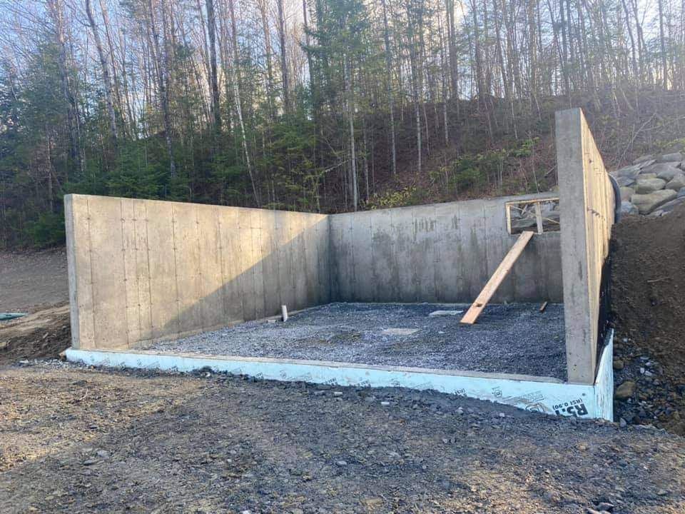 Murs de fondation en béton sur une base de gravier, adossés à un flanc de colline boisé.