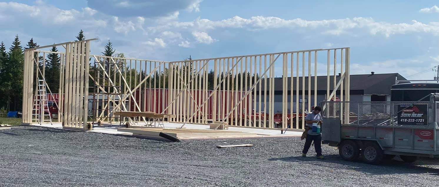 La charpente en bois d'un bâtiment en construction, avec une personne debout près d'une remorque sur un terrain gravillonné.