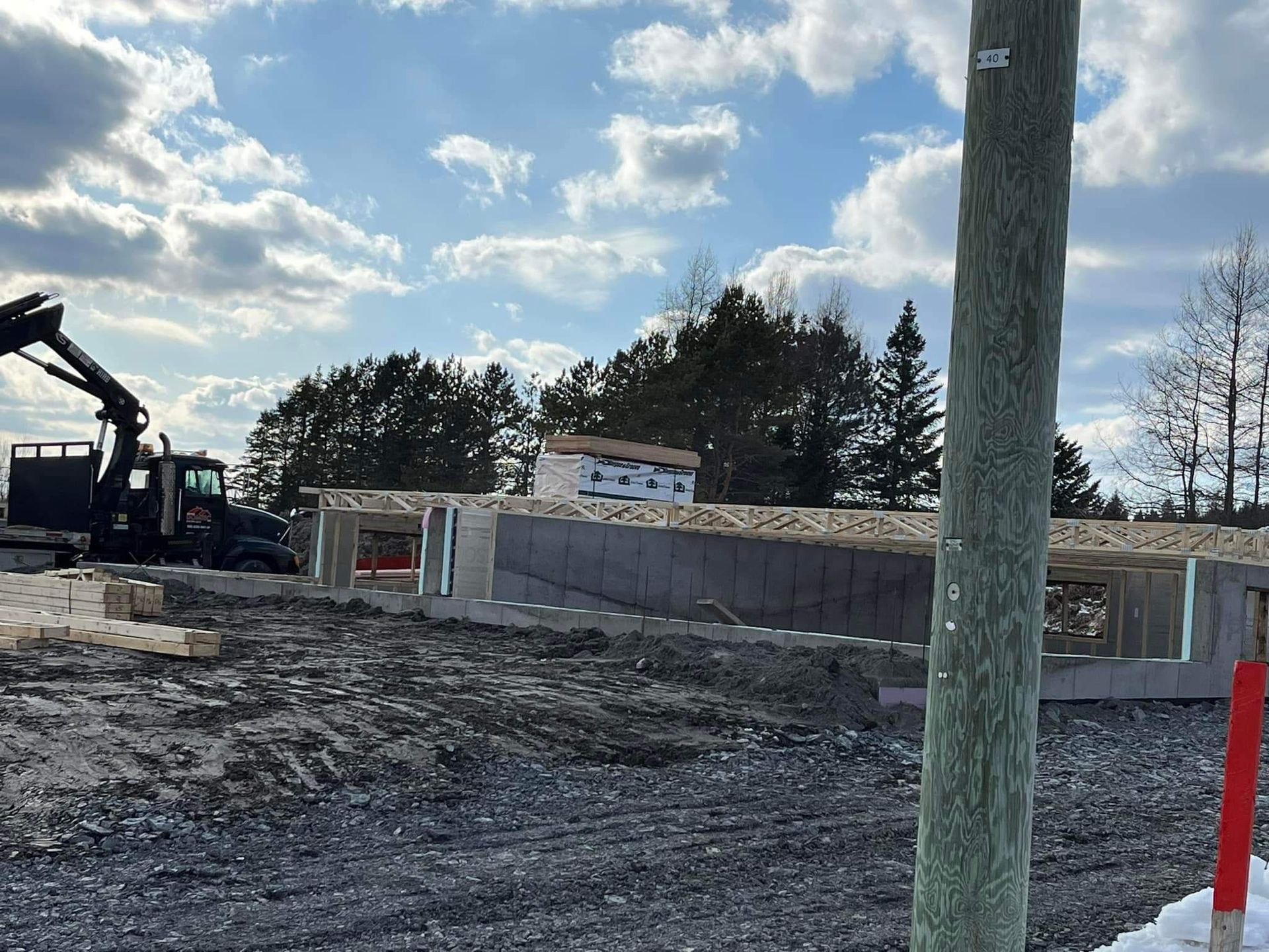 Chantier de construction avec une structure en béton partiellement construite, une excavatrice et un poteau électrique sur fond de ciel nuageux.