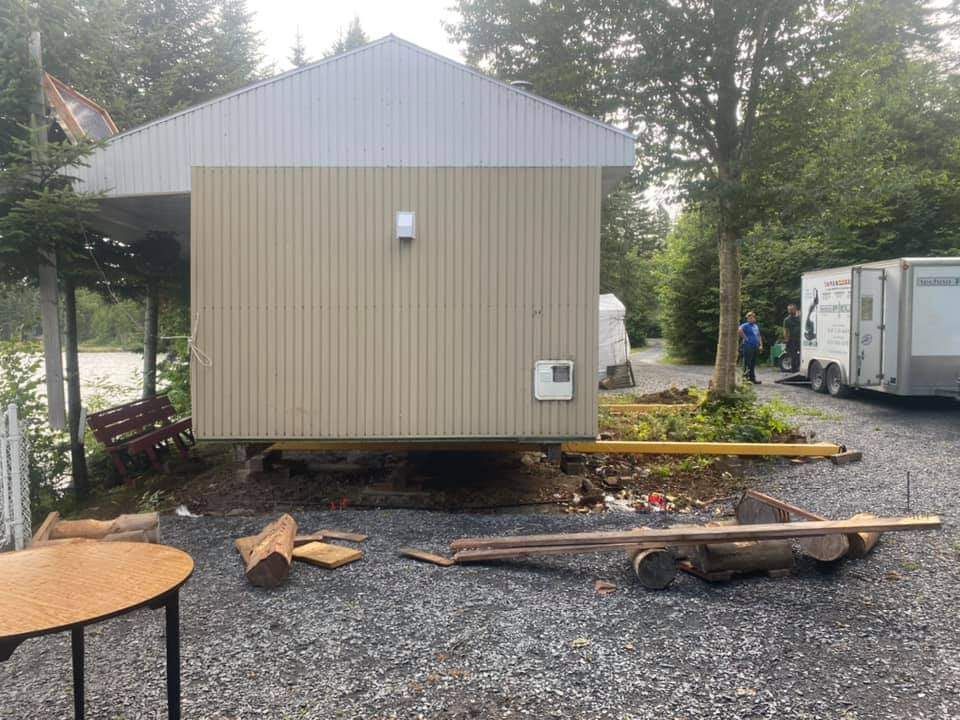 Un bâtiment beige est déplacé par des engins de chantier, avec des grumes et une charrette devant lui. Une personne se tient à proximité.