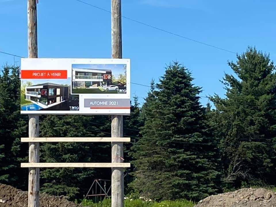 Panneau publicitaire pour une maison moderne en construction, fixé sur des poteaux en bois, avec des arbres verts en arrière-plan.