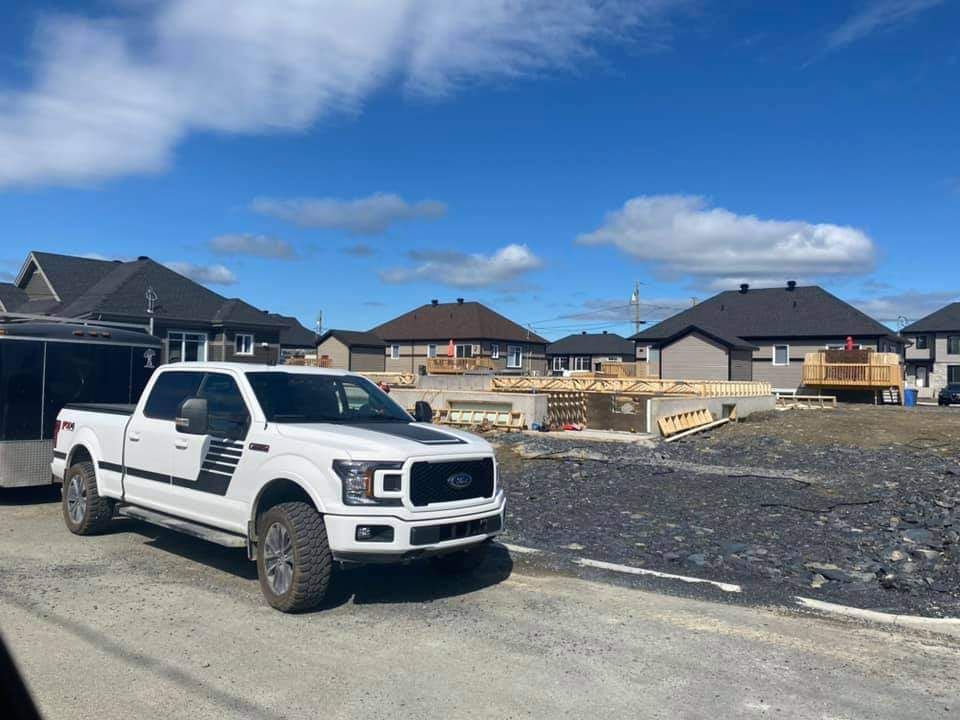 Un pick-up Ford blanc avec remorque est stationné sur un chantier de construction de maisons par une journée ensoleillée.
