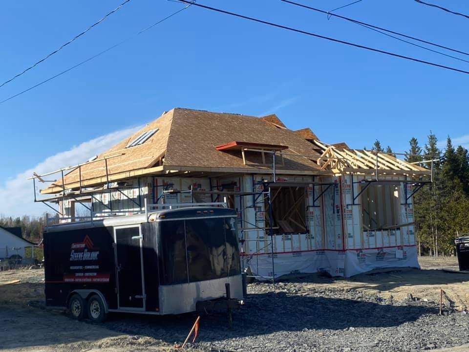 Maison en construction avec charpente apparente et toiture partiellement recouverte de bardeaux. Une caravane est garée devant.