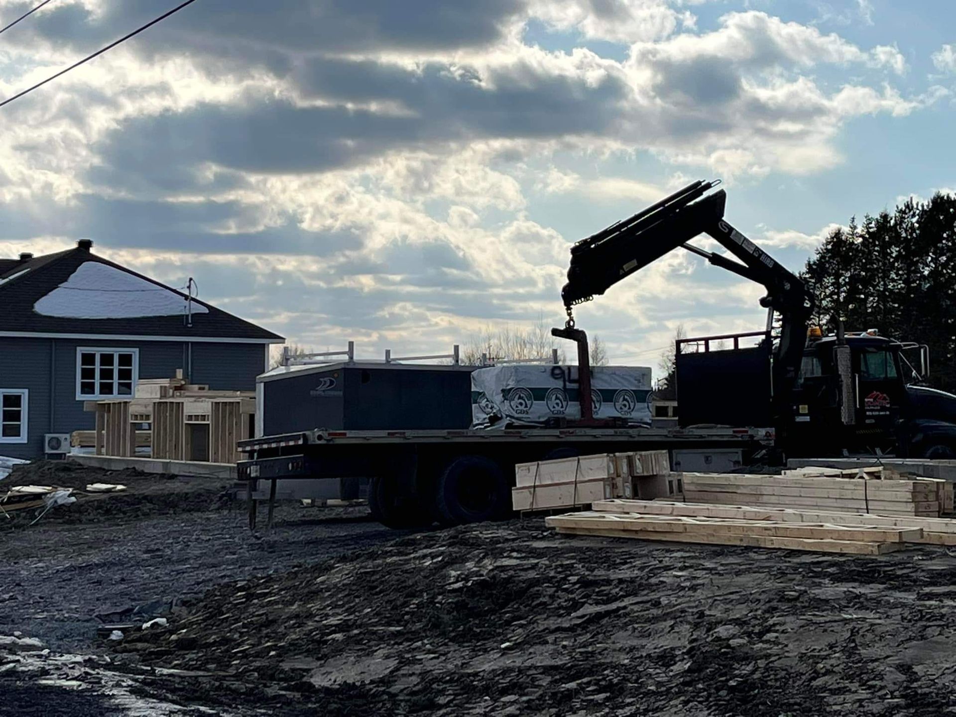 Une grue charge du bois sur un chantier de construction près d'une maison bleue, sous un ciel nuageux.