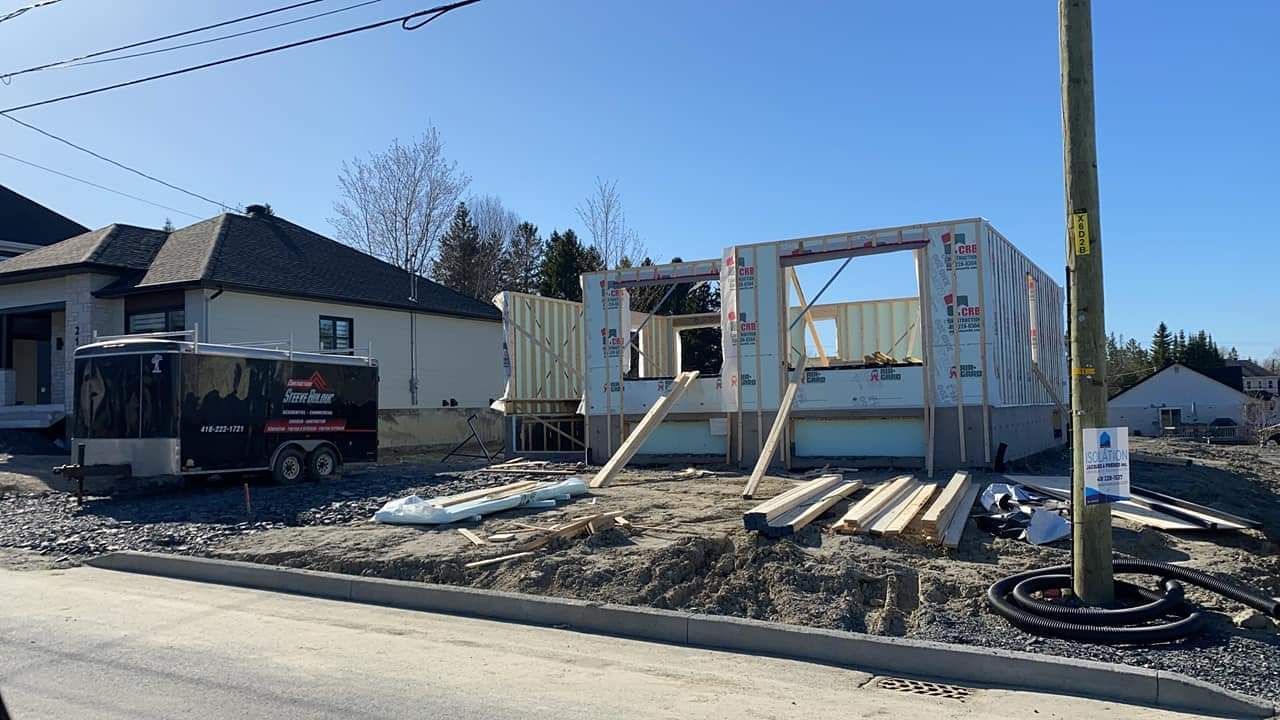 Chantier de construction : Ossature d'une nouvelle maison à côté d'une maison existante, journée ensoleillée.