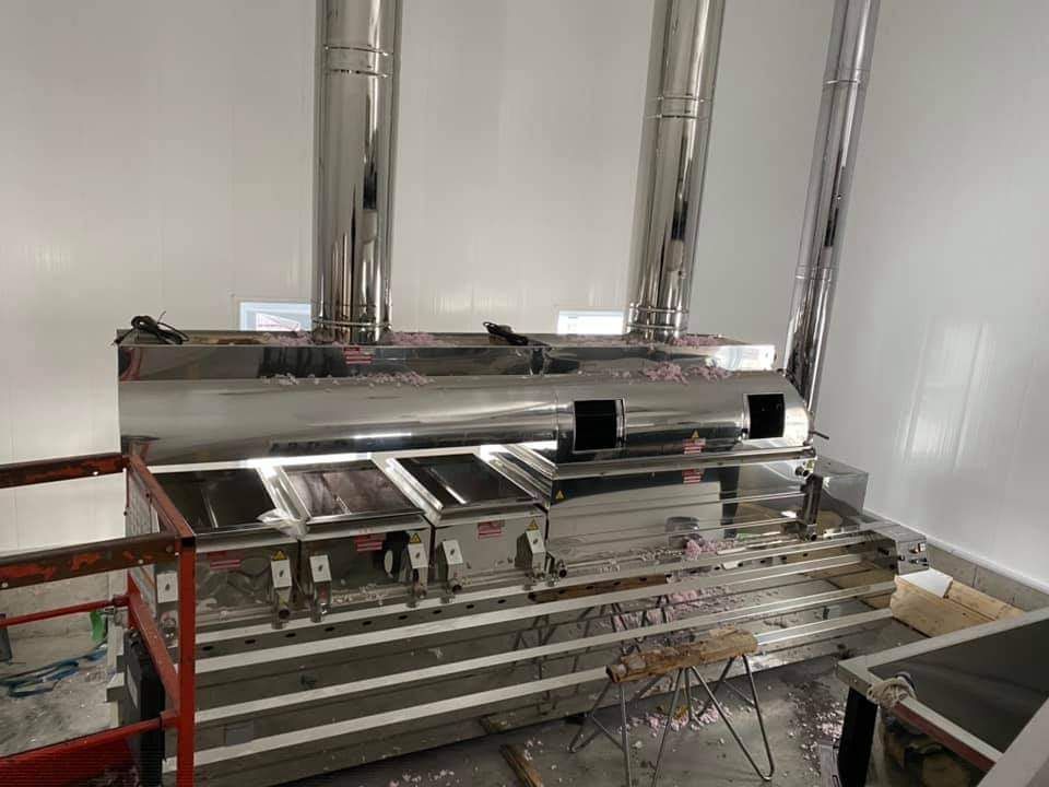 Grand évaporateur à sirop d'érable en acier inoxydable avec trois cheminées dans une pièce aux murs blancs.