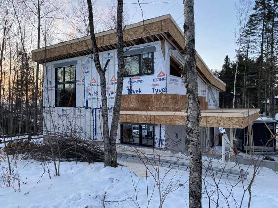 Maison en construction dans une forêt enneigée ; toit en panneaux OSB et bâche Tyvek visibles.