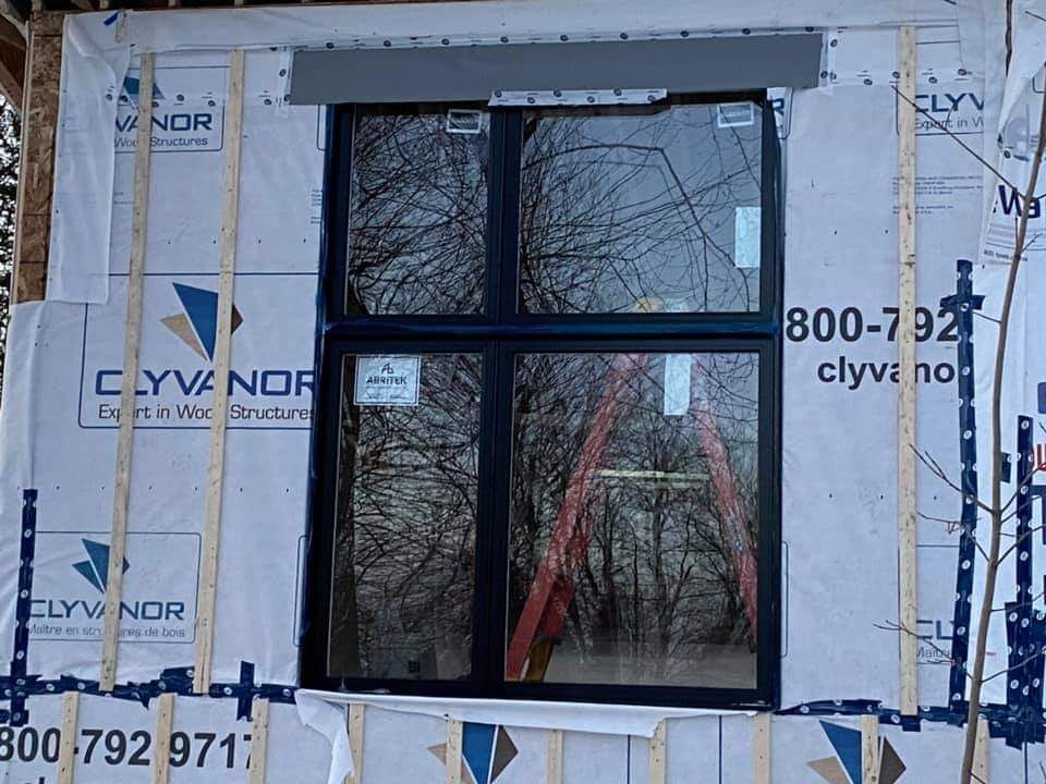 Fenêtre installée dans un cadre de bâtiment, recouverte d'un revêtement étanche blanc. Cadre de fenêtre noir, échelle visible dans le reflet.