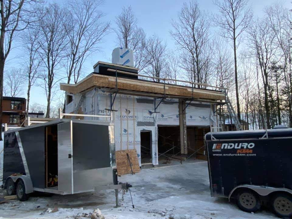 Chantier de construction : bâtiment partiellement construit avec charpente apparente, remorques stationnées à l'extérieur, paysage hivernal.