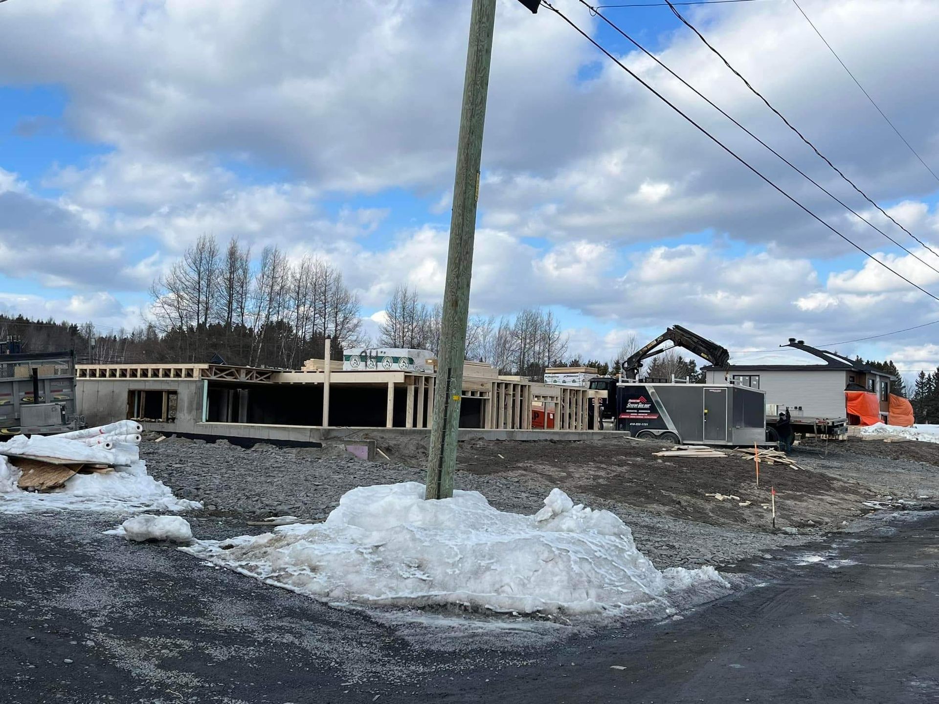 Chantier : charpente en bois, gravier, neige, poteau électrique, ciel couvert.