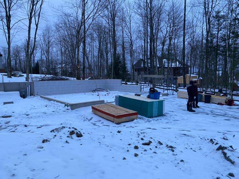 Chantier de construction sous la neige ; fondations, bois d’œuvre et deux personnes. Arbres dénudés en arrière-plan.