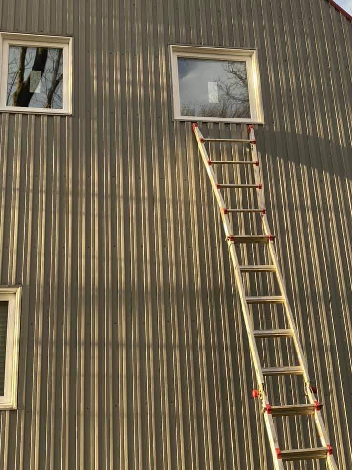 Une échelle appuyée contre un bâtiment gris atteint une fenêtre. La lumière du soleil projette des ombres.