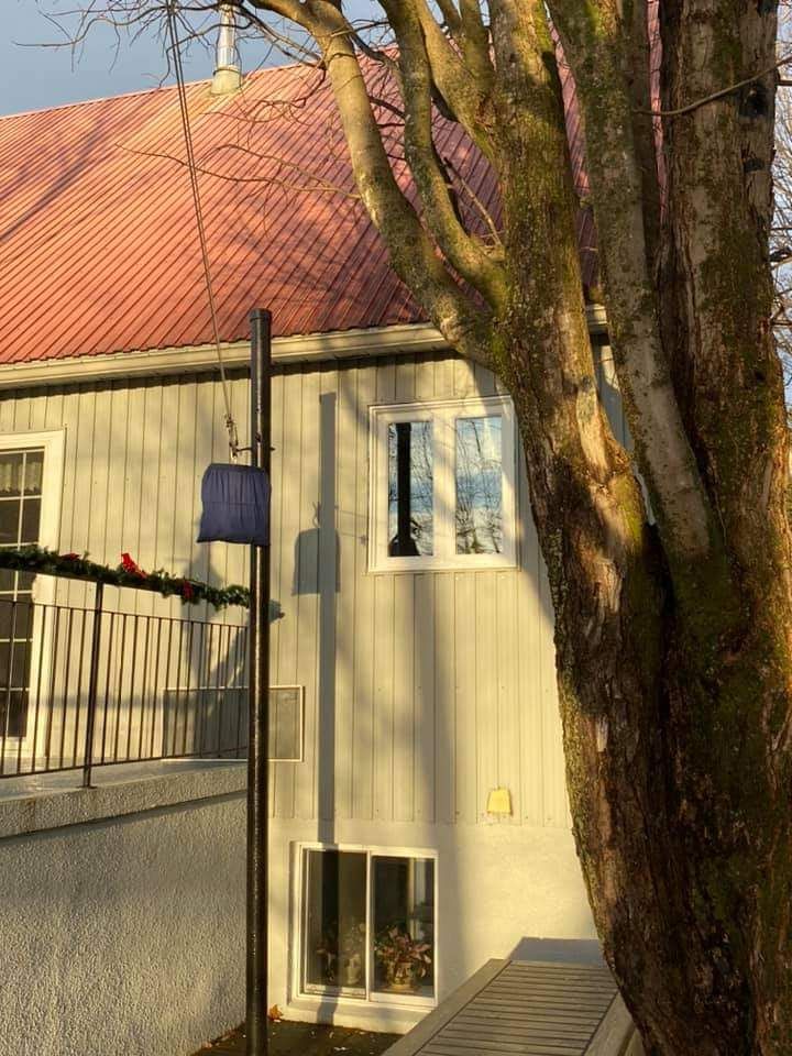 Bâtiment aux façades grises et au toit rouge, arbre au premier plan. Fenêtre et ombres projetées par un poteau métallique et un arbre.