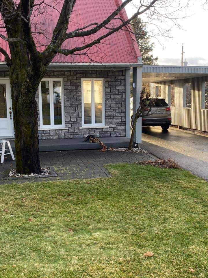 Une maison en pierre au toit rouge, deux fenêtres et une voiture sous un abri.