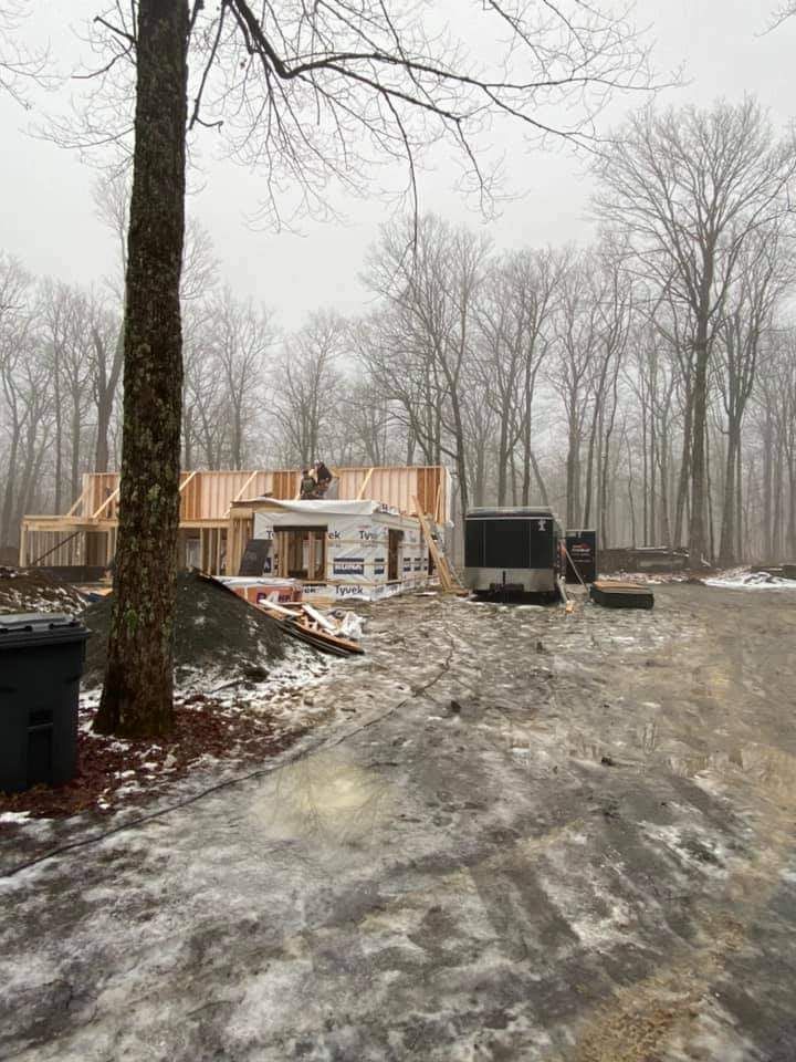 Chantier de construction en hiver, avec une maison à ossature partiellement visible, entourée d'arbres et d'un chemin boueux.