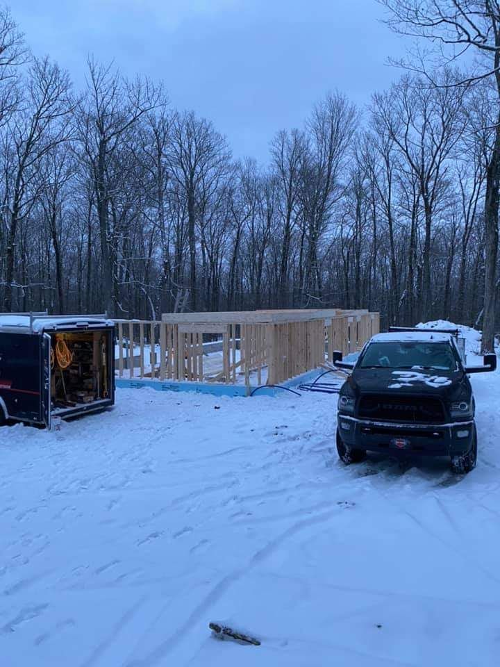 Chantier de construction en hiver, avec une charpente en bois inachevée, entouré de neige, une remorque et un camion.