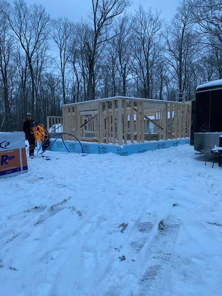 Charpente d'un petit bâtiment en construction, sous la neige. Deux personnes travaillent à l'extérieur. Arbres en arrière-plan.