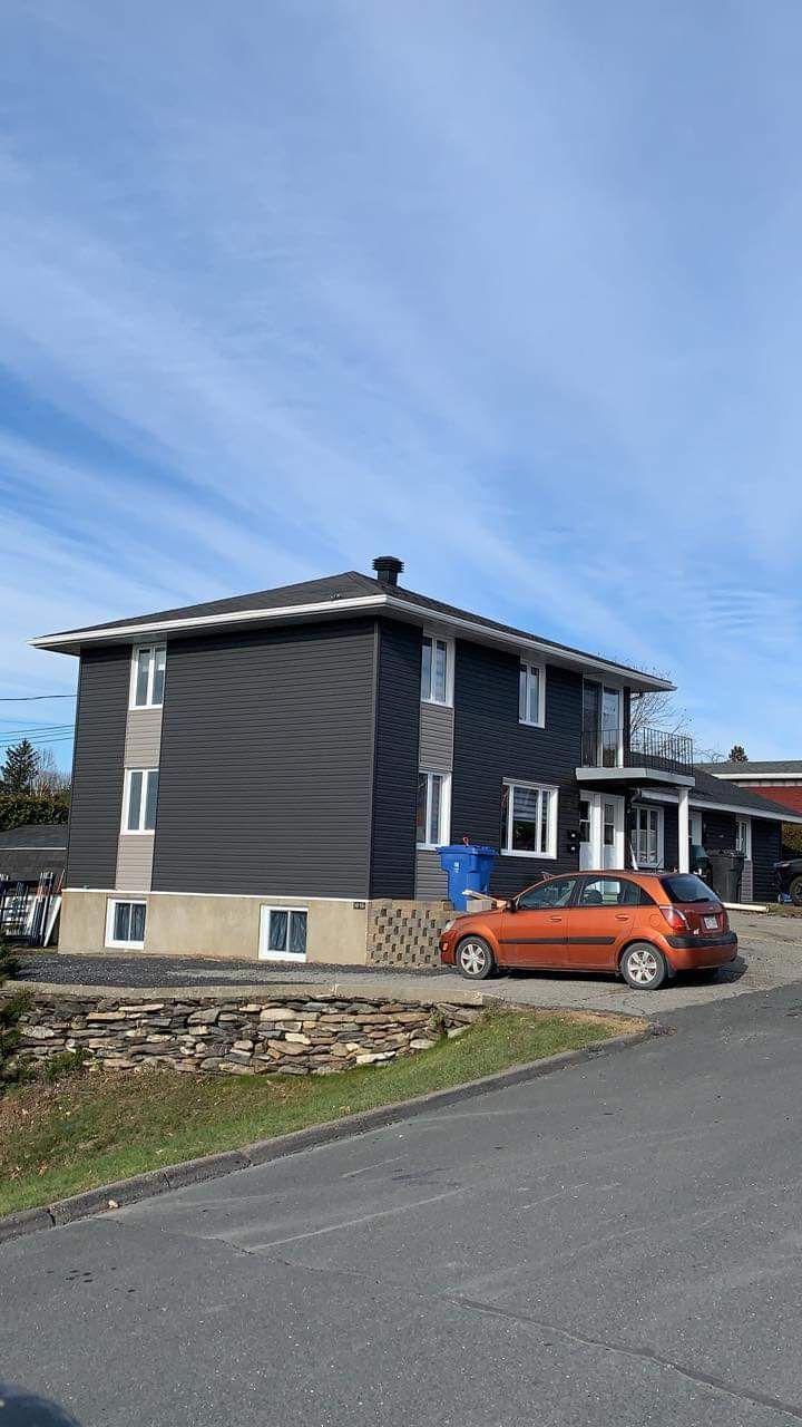 Immeuble gris foncé à plusieurs étages avec des touches de beige, voiture orange garée devant, ciel bleu.