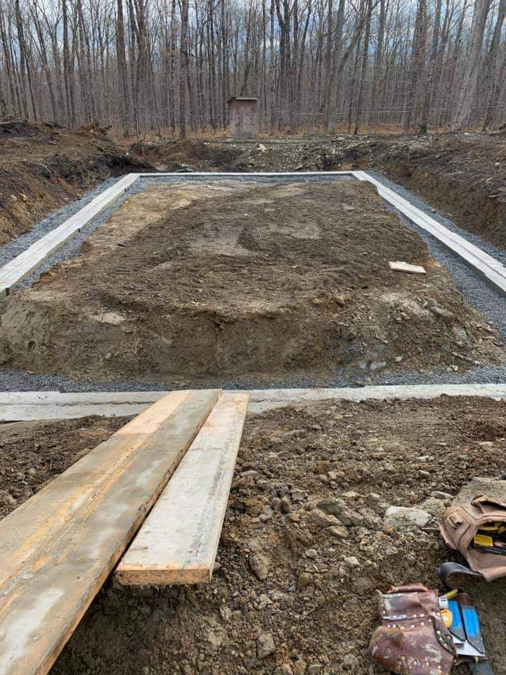 Chantier : fondations rectangulaires, périmètre de gravier, monticule de terre, bois d’œuvre, outils. Forêt en arrière-plan.