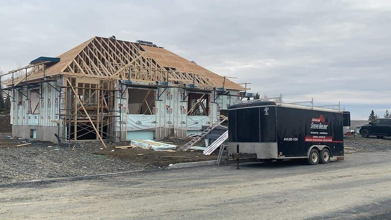 Construction d'une maison : ossature bois, partiellement recouverte d'isolant. Une remorque noire est garée à côté.
