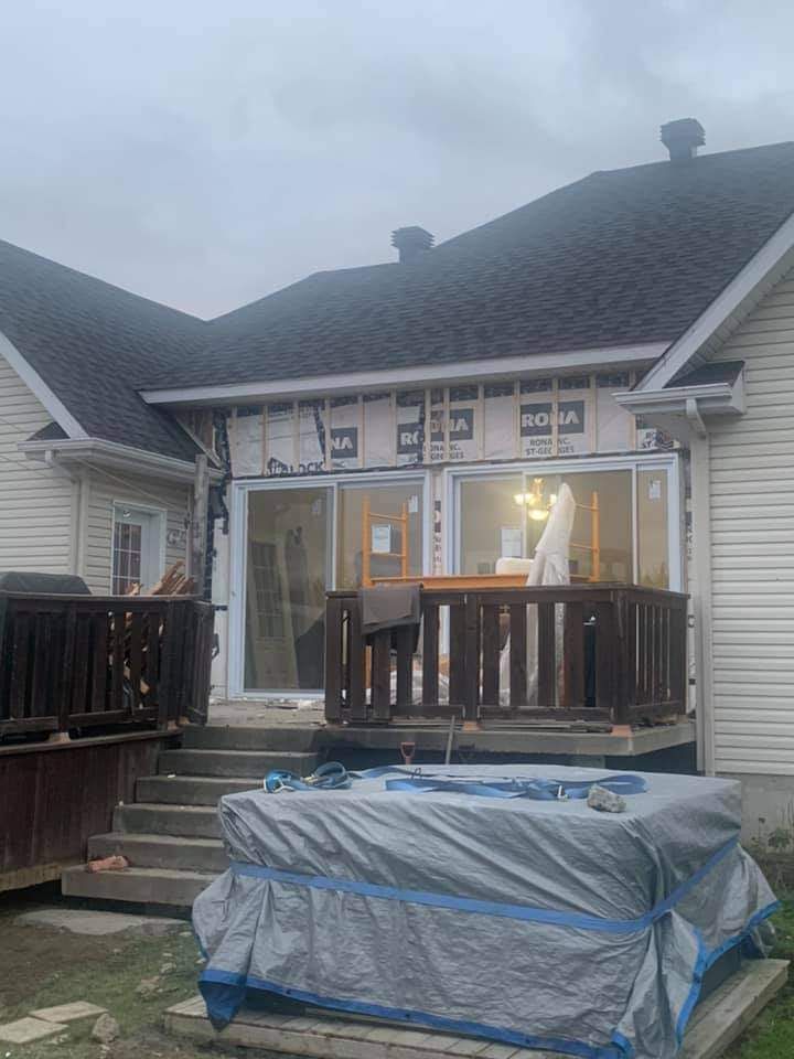 Arrière d'une maison en construction avec terrasse, escalier et objets recouverts ; ciel gris.