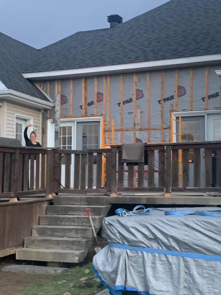 Une femme fait signe depuis une terrasse. Travaux de construction sur la façade d'une maison ; charpente apparente. Une bâche grise recouvre quelque chose sous la terrasse.