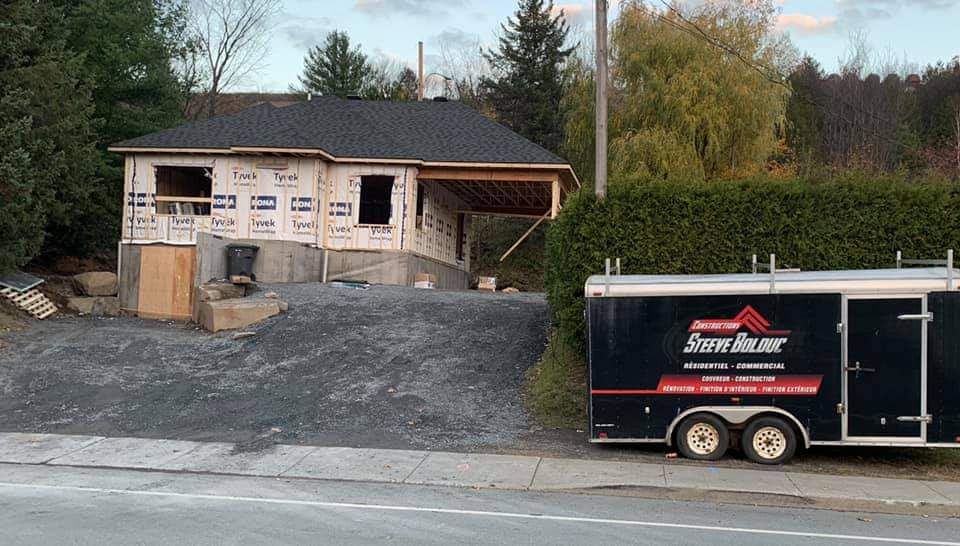 Chantier de construction avec une maison partiellement construite, une caravane et une allée en gravier.