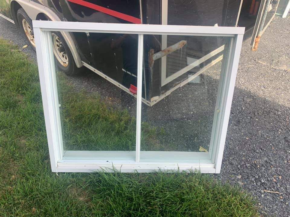 Fenêtre blanche à deux vantaux, posée sur l'herbe. Une caravane est visible en arrière-plan.