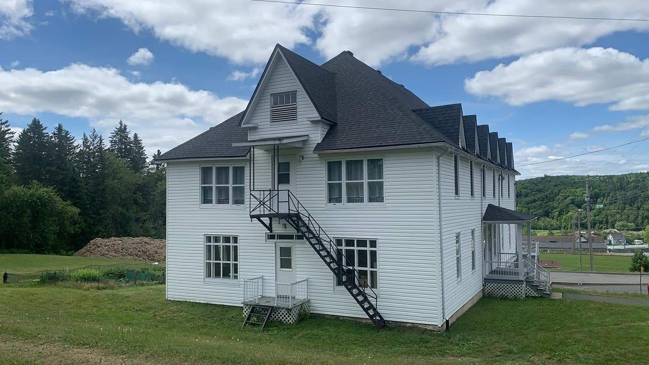 Bâtiment blanc à plusieurs étages, avec toit noir et escalier de secours, sur une colline herbeuse sous un ciel nuageux.