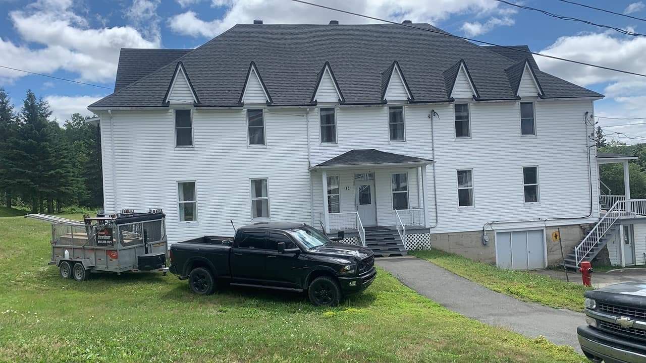Maison blanche à deux étages avec toit noir, camionnette noire et remorque garées devant sur une pelouse.
