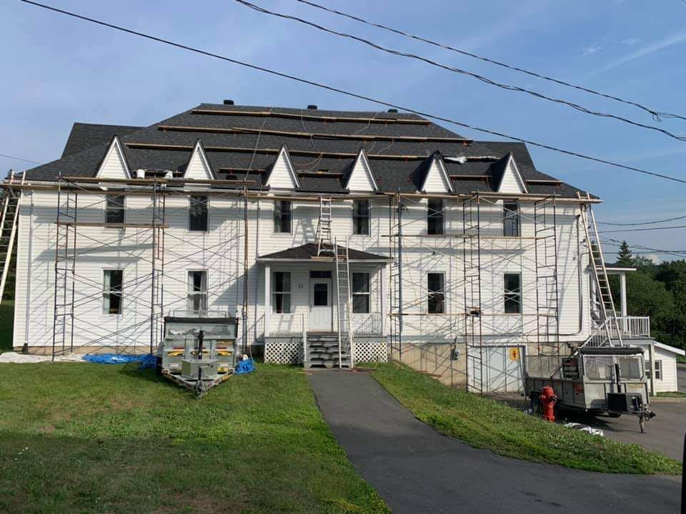 Bâtiment blanc avec échafaudages, toiture partiellement refaite, sur une colline herbeuse ; ciel bleu.