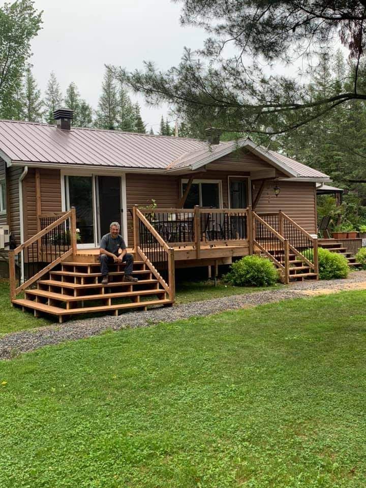 Un homme est assis sur les marches en bois d'une cabane au toit et à la terrasse bruns. Une pelouse verte l'entoure.