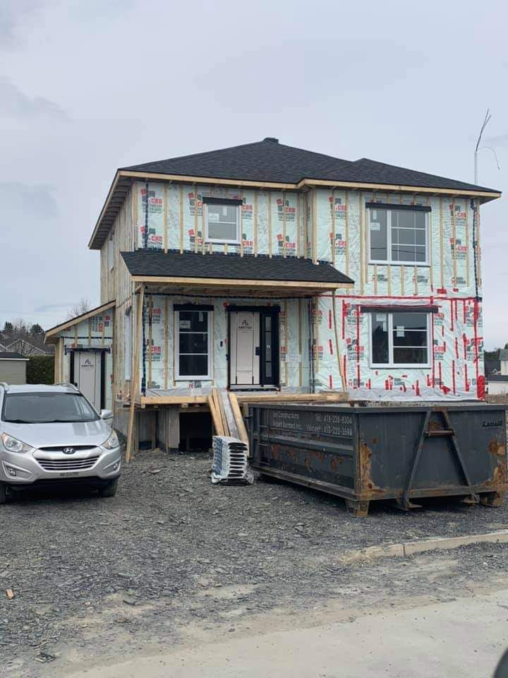 Maison de deux étages en construction, avec charpente apparente, bardage et toit noir. Une voiture argentée est garée devant.