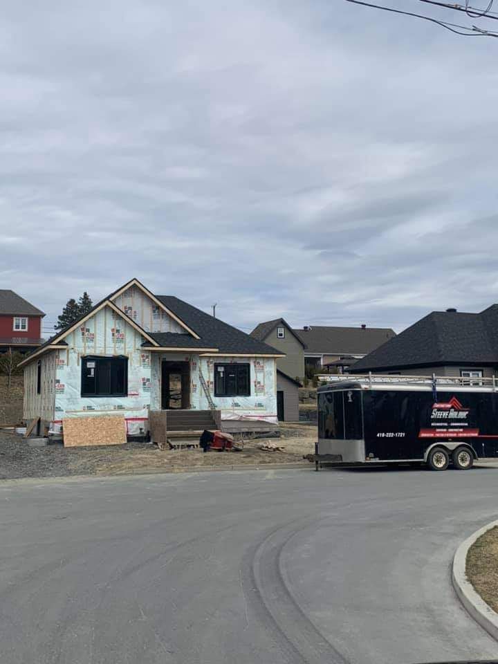 Maison en construction avec toit et bardage noirs. Une caravane est stationnée à proximité, dans une rue pavée.