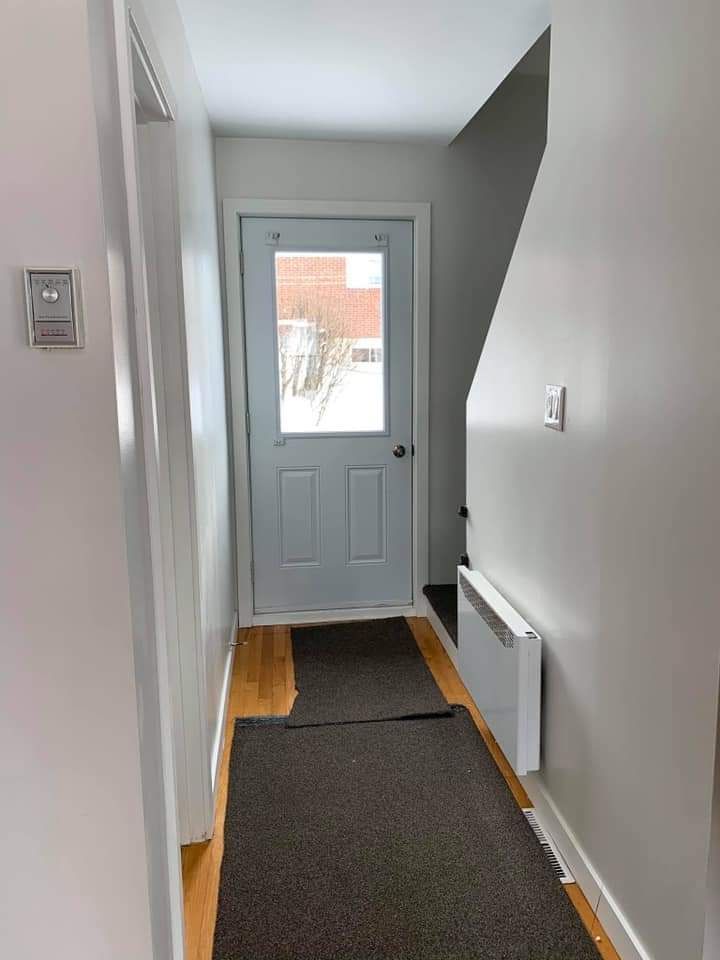Couloir étroit avec une porte au fond, deux tapis sombres au sol, des murs blancs et un escalier.