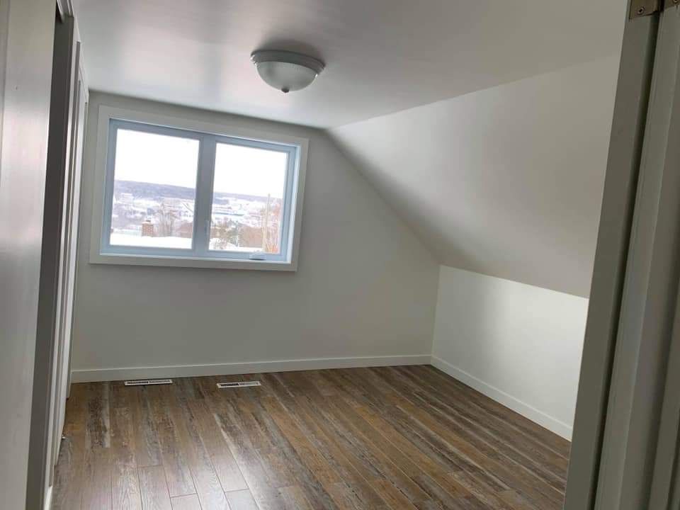 Pièce vide avec plafond mansardé, fenêtre et sol imitation bois. Murs et plafond blancs.