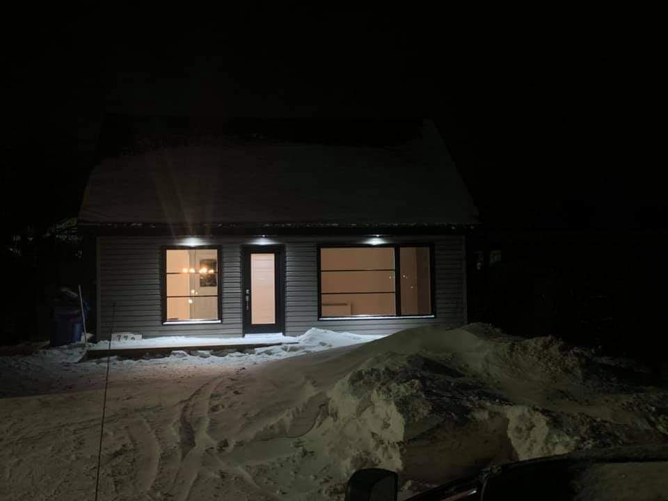 Petit bâtiment enneigé la nuit. Fenêtres et éclairage extérieur éclairés. Sol enneigé. Ciel sombre.