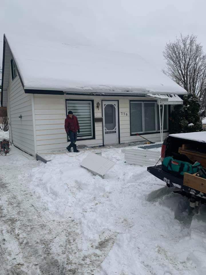 Un homme se tient devant une maison blanche recouverte de neige. Un camion transportant des outils est stationné dans la neige.