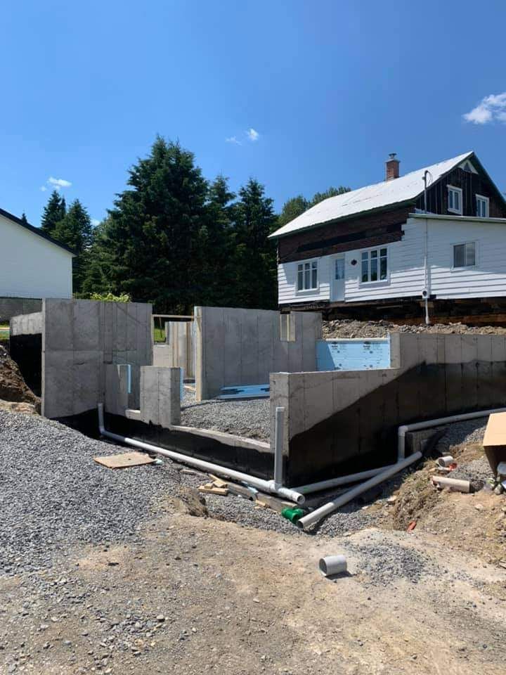 Murs de fondation d'une maison en construction ; gravier, tuyaux et maison en arrière-plan.