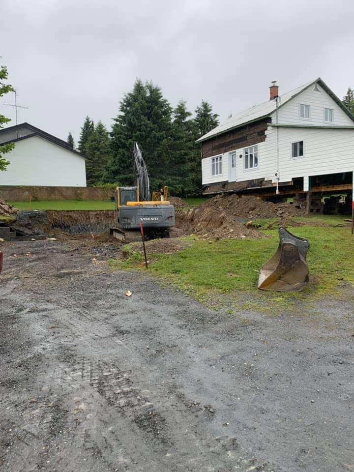 Une pelleteuse effectue des travaux de terrassement près d'une maison, probablement pour des travaux de construction ou de fondation, sur un terrain gravillonné. Ciel couvert.
