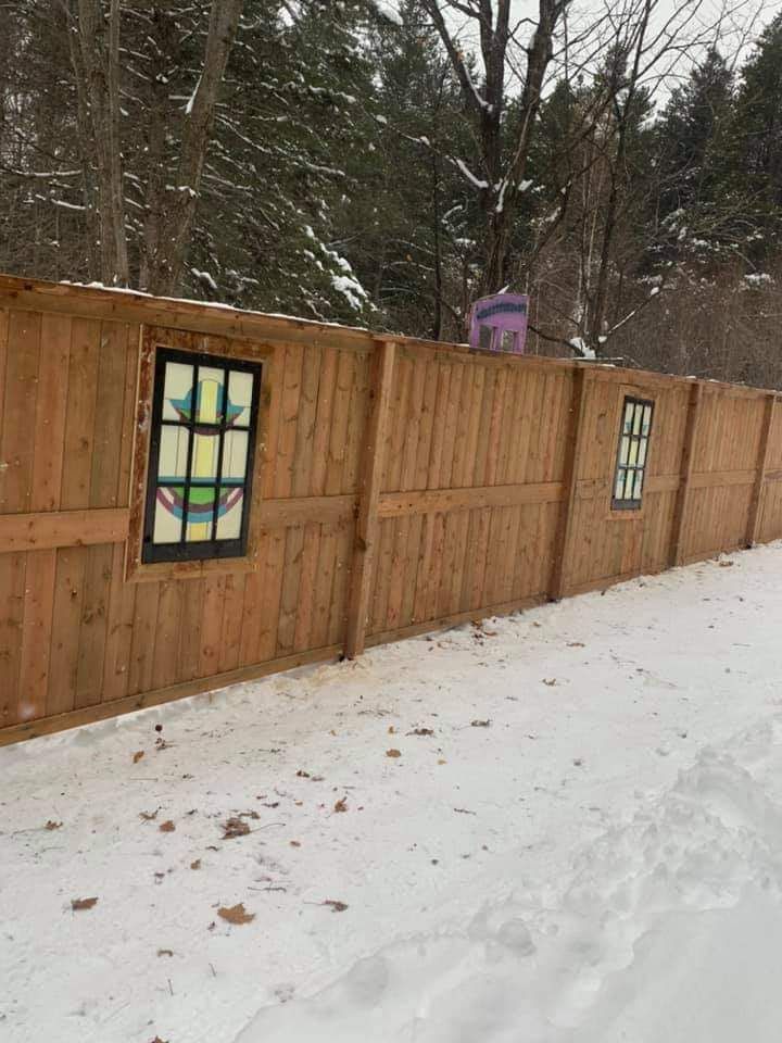 Clôture en bois ornée de vitraux dans une zone enneigée, avec des arbres en arrière-plan.