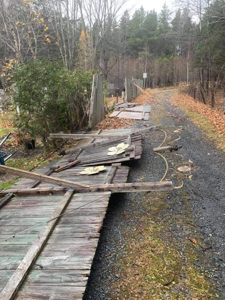 Passerelle en bois délabrée longeant un chemin de gravier dans une zone boisée. Des débris et des feuilles mortes sont visibles.